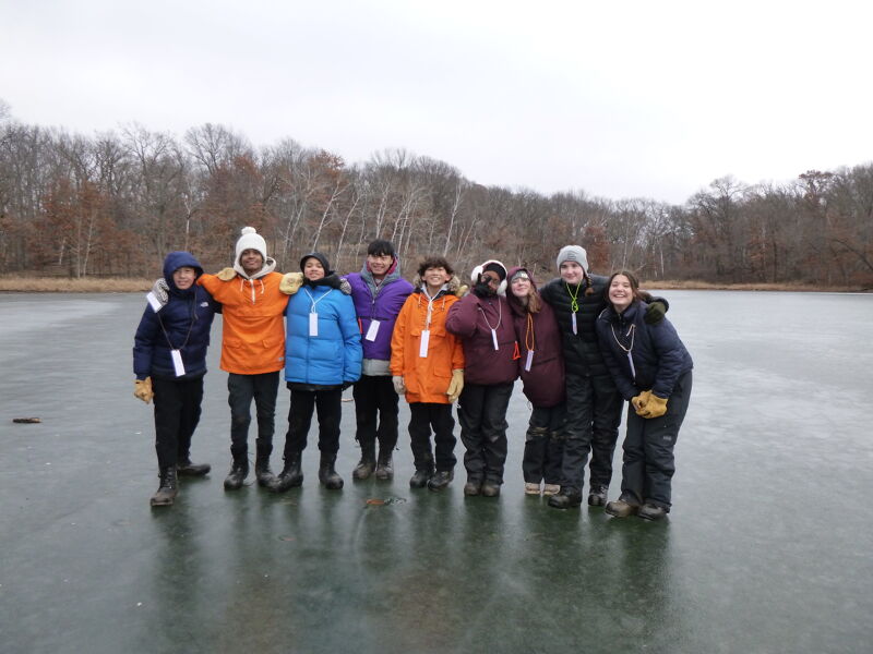 The image shows a group of people standing on a frozen lake. They are all wearing winter coats and appear to be enjoying the outdoors. The background features trees and a cloudy sky, suggesting a cold day. The people are smiling and seem to be having a good time together.